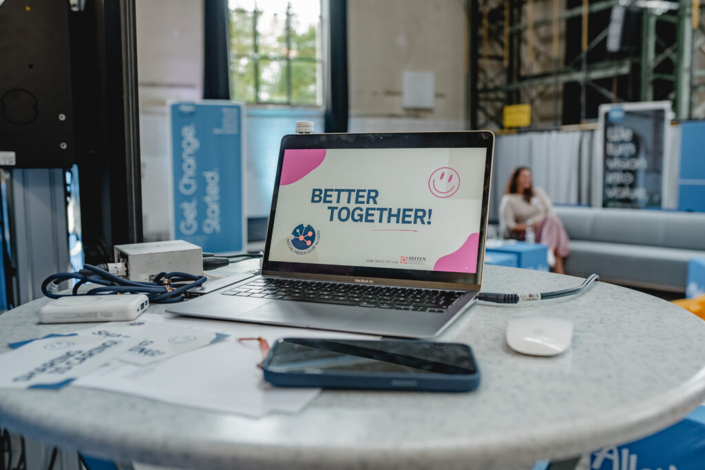 Zu Sehen ist ein Laptop auf einem runden Stehtisch in einem Veranstaltungsraum. Der Laptop ist geöffnet und auf dem Bildschirm steht "Better Together".